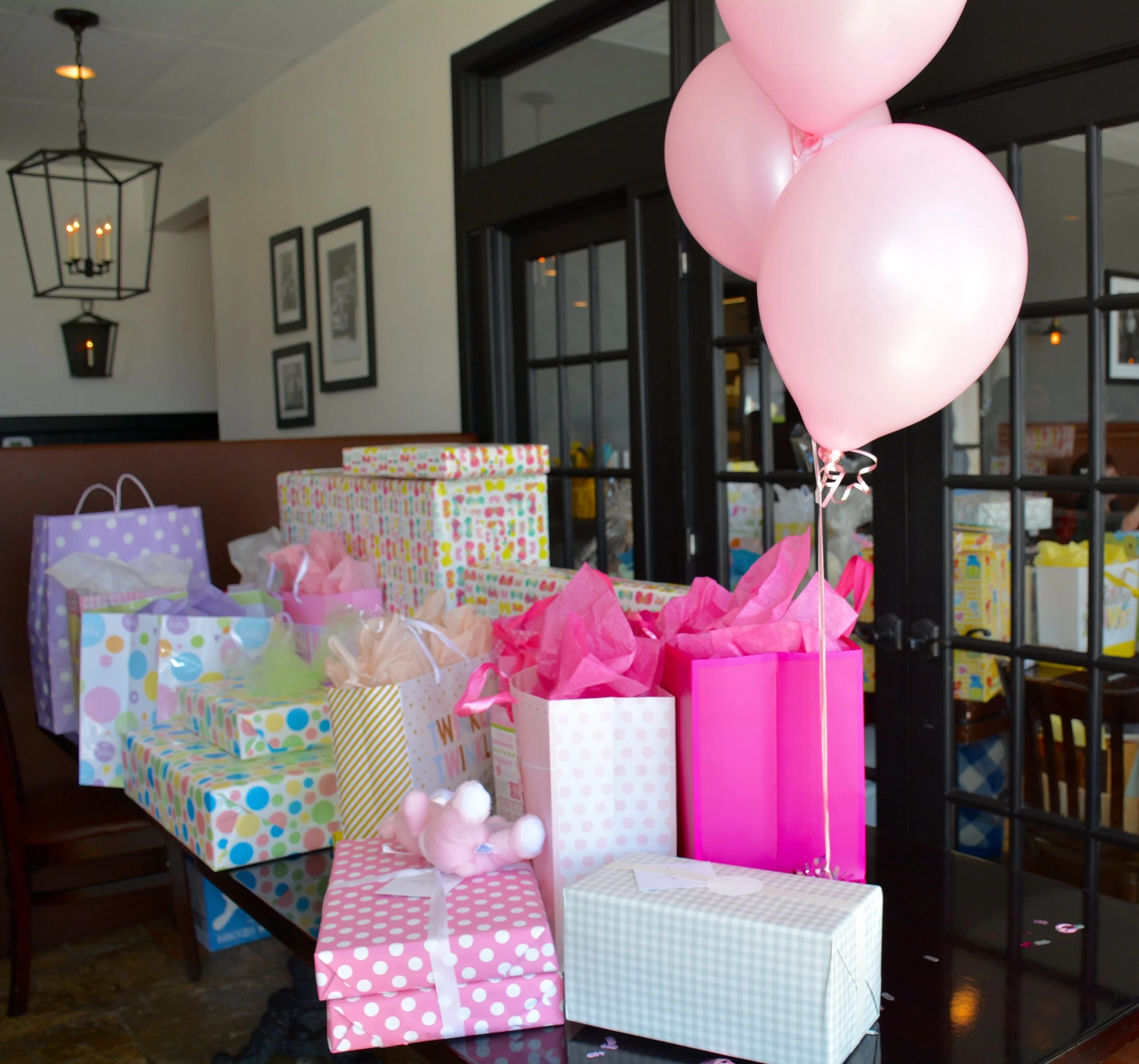 A table filled with colorful wrapped gifts and gift bags, decorated with pink balloons, in a warmly lit room with picture frames on the wall