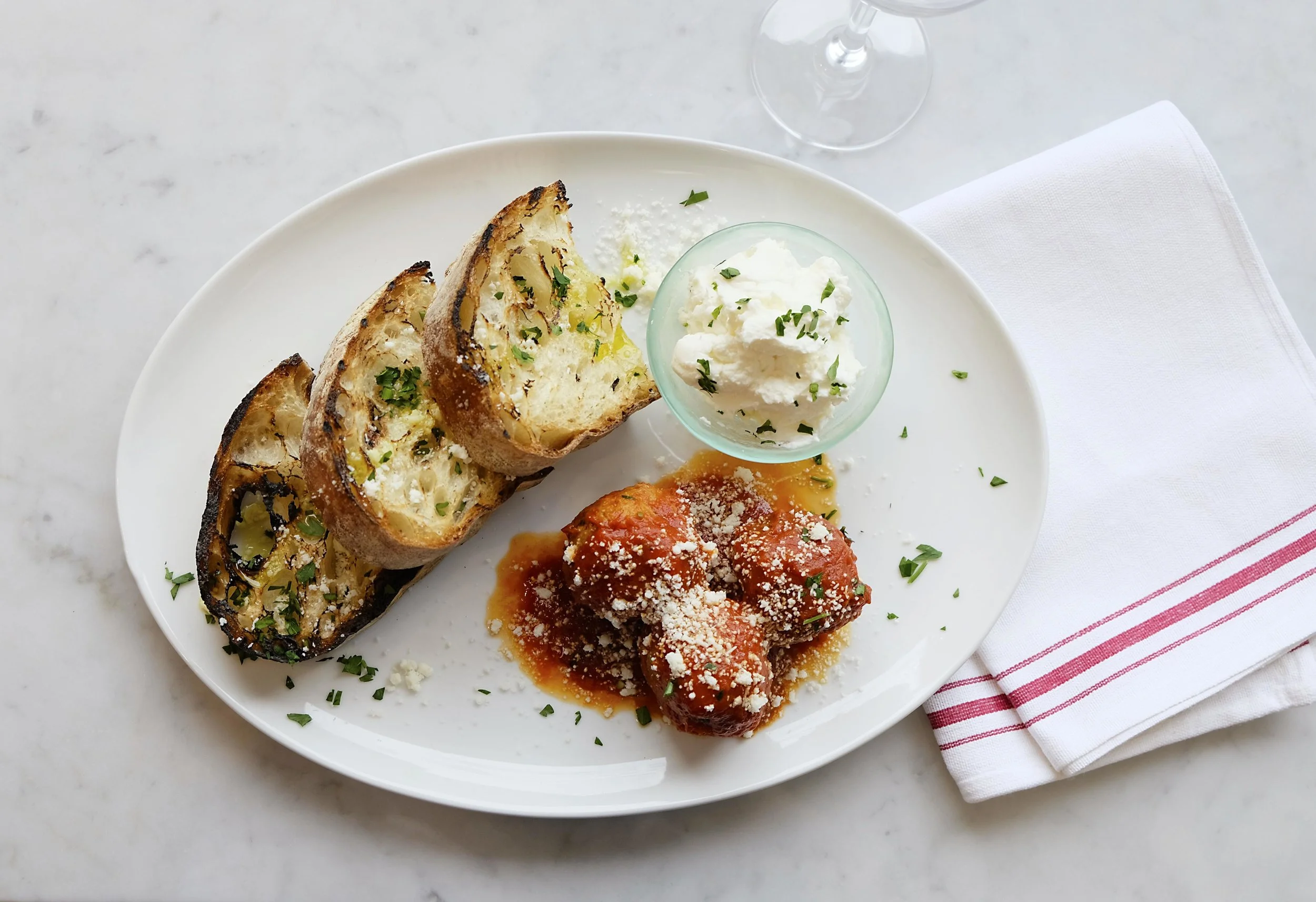 Plate of toasted bread, meatballs in sauce topped with cheese, and a small bowl of whipped cheese on a marble table, warm earthy tones